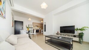 Un salon moderne avec un canapé, une table basse, un téléviseur et une cuisine en arrière-plan.