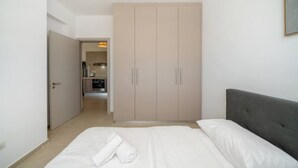 Une chambre à coucher comprenant un lit, une tête de lit grise, une armoire et un couloir menant à une petite cuisine.