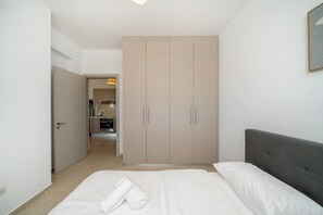 Un dormitorio con cama, cabecera gris, armario y un pasillo que da a una kitchenette.