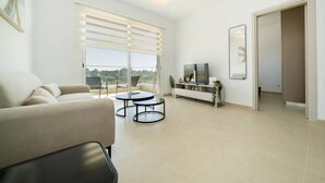 A modern living room with a sofa, a coffee table, a TV on a stand, and a balcony with a view.