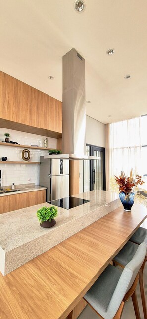 A modern kitchen with a central island, stainless steel appliances, wooden cabinetry, and a large window providing natural light. - PARQUE PRADO IMÓVEIS SIRIUS SKY APTO 807 (Campinas)