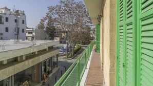 Varanda com persianas verdes dando para uma rua com edifícios e árvores.