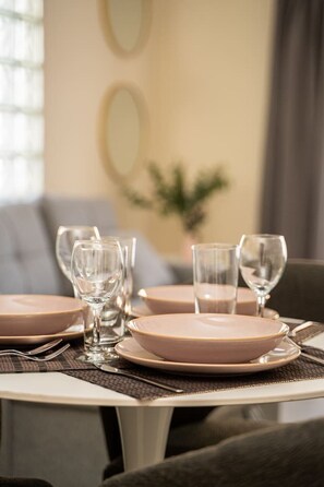 Une table à manger dressée avec des assiettes roses et des verres transparents, avec un canapé et une plante en arrière-plan.