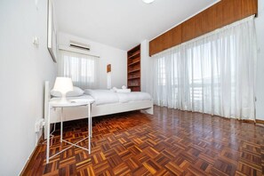 Quarto com cama, mesa de cabeceira, abajur, piso de madeira, janela com cortinas leves e estante embutida.