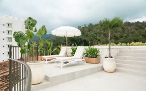 Eine Dachterrasse mit weißen Liegestühlen, einem weißen Sonnenschirm und Topfpflanzen.