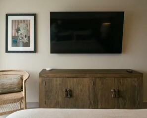 Ein Zimmer mit einem großen Flachbildfernseher an der Wand, einem Holfach darunter und einem Korbstuhl links.