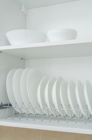 Une cuisine avec des meubles blancs, un présentoir métallique contenant des assiettes blanches et un grand saladier blanc sur l’étagère supérieure.