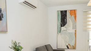 Ett modernt vardagsrum med en grå soffa, ett glasbord med vita stolar, en stor abstrakt målning och en luftkonditioneringsanläggning.
