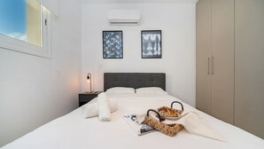 En pænt redt seng med en kurv med mad, et magasin og et håndklæde på et hvidt sengetæppe. Der er et natbord med en lampe og et billede i ramme på væggen.