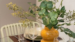 Et spisebord med glasplade, omgivet af stole, med en vase med grønt og en gul vase.