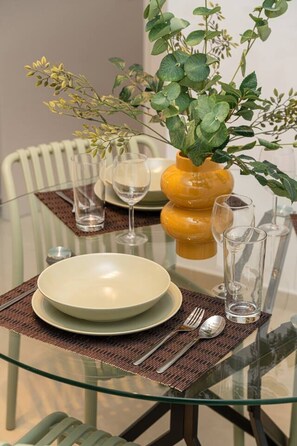 A dining table with a glass top, surrounded by chairs, with a vase of green foliage and a yellow vase.