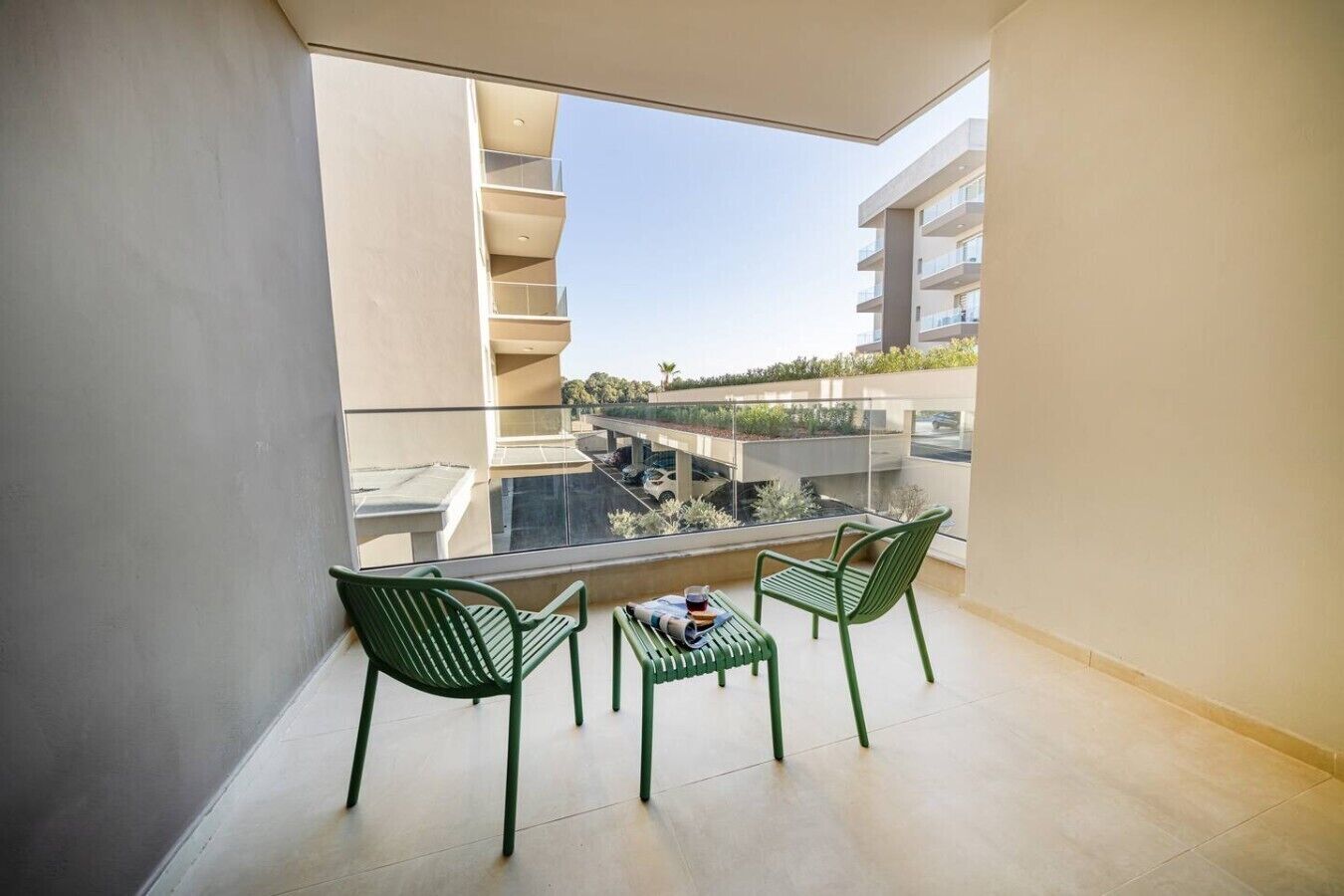 Ein Balkon mit zwei grünen Stühlen, einem kleinen Tisch und freiem Blick auf angrenzende Gebäude und einen Parkplatz.