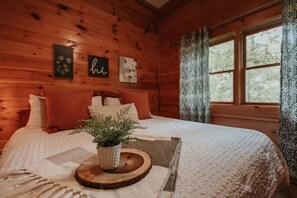 Ein Schlafzimmer einer Holzhütte mit einem Bett, Kissen, einer Pflanze und einem Fenster mit Vorhängen.