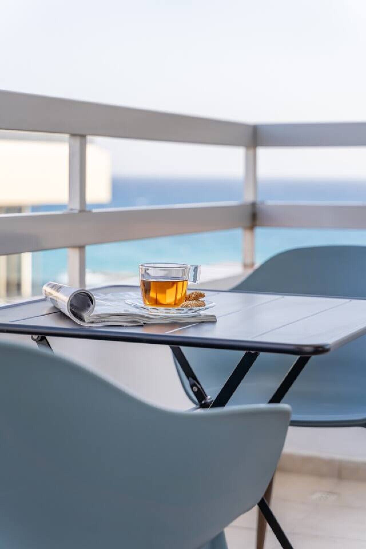 Una mesa con una taza de té, un periódico y un teléfono inteligente en un balcón con vistas al mar.