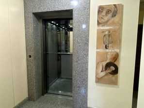 Un pasillo con un ascensor de cristal, una obra de arte en la pared y una puerta con interruptor de luz.