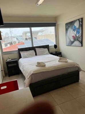 Un dormitorio con cama, mesita de noche, ventana con vistas a edificios y un cuadro en la pared.