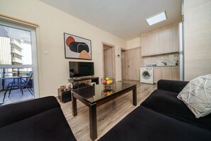 Ein modernes Wohnzimmer mit Flachbildfernseher, einem Esstisch mit Obst, einer Couch und einer Küchenzeile mit Waschmaschine.