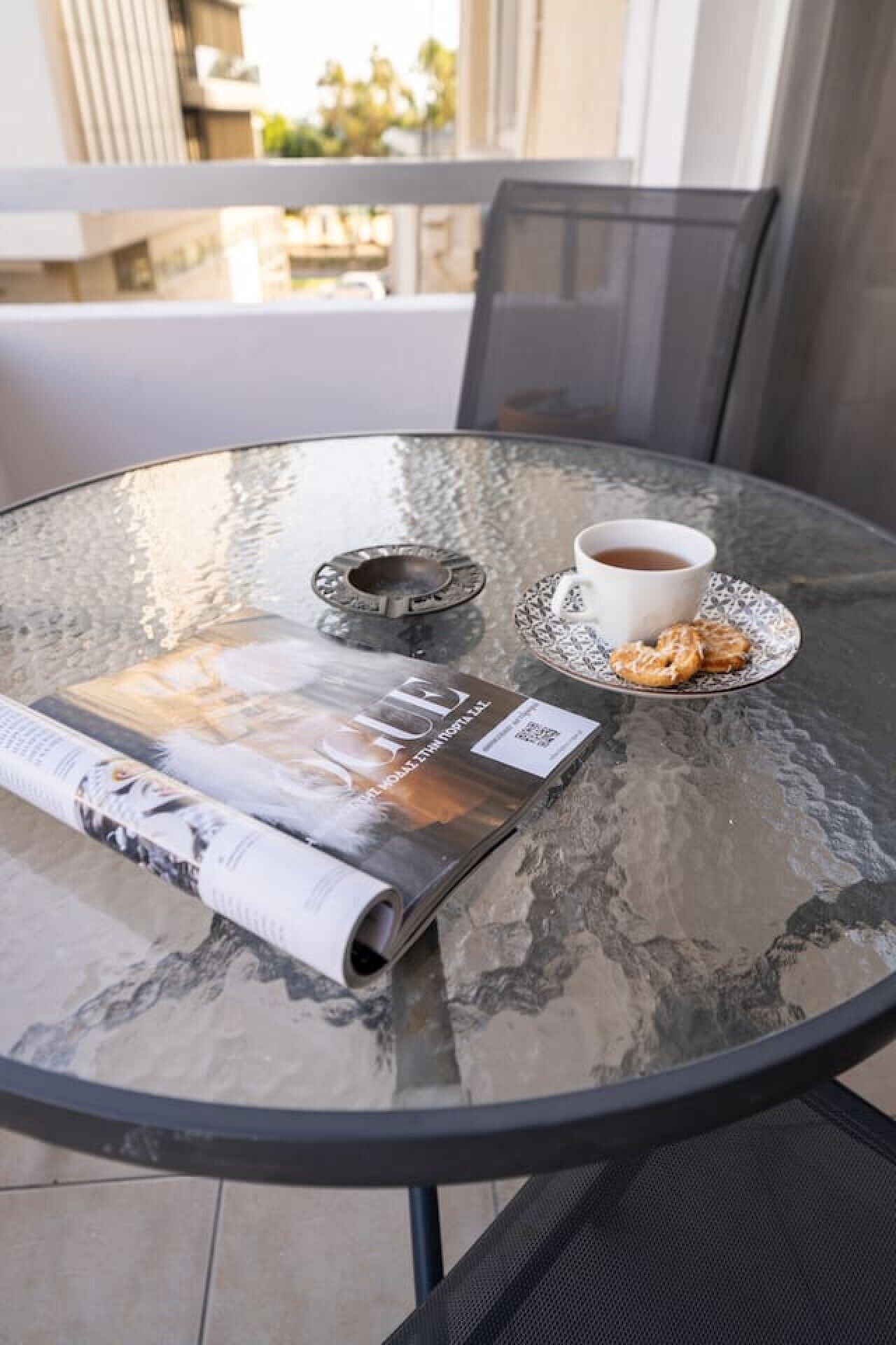 Et rundt glastil med et magasin, en kop kaffe og en tallerken med småkager på en balkon.