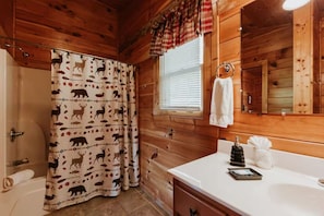 Ein Badezimmer einer Holzhütte mit Duschvorhang mit Tiermotiven, einem weißen Waschbecken mit Seifenspender und einem Fenster mit Jalousien.