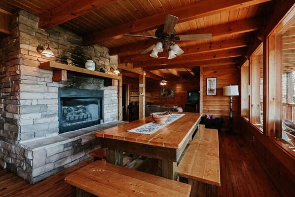 A rustic cabin interior with a stone fireplace, wooden beams, a ceiling fan, and a large window. - 6BR/6BA Mountain Lodge with Perfect Views (Gatlinburg)