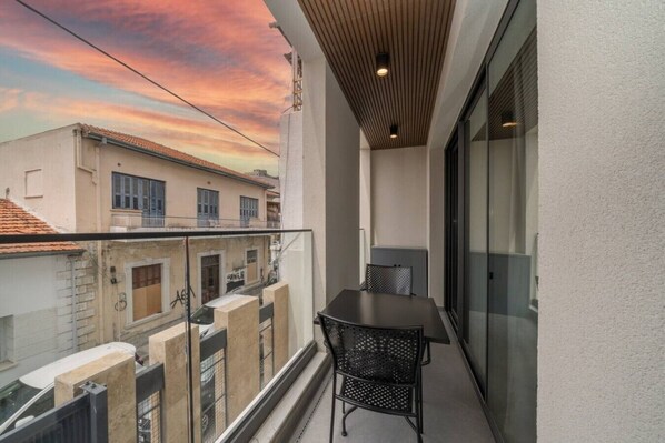 Un balcón con mesa y sillas, con vistas a un paisaje urbano que incluye edificios y un atardecer.