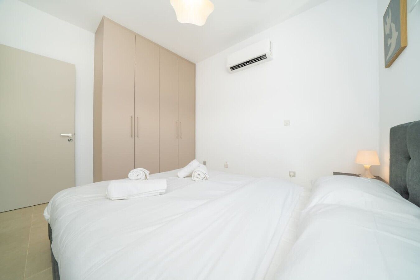 A bedroom with a bed, a wardrobe, a bedside table with a lamp, and an air conditioning unit.