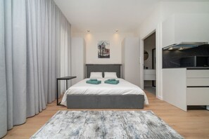 Een moderne hotelkamer met een groot bed, een klein nachtkastje, een bureau met stoel en een kitchenette.
