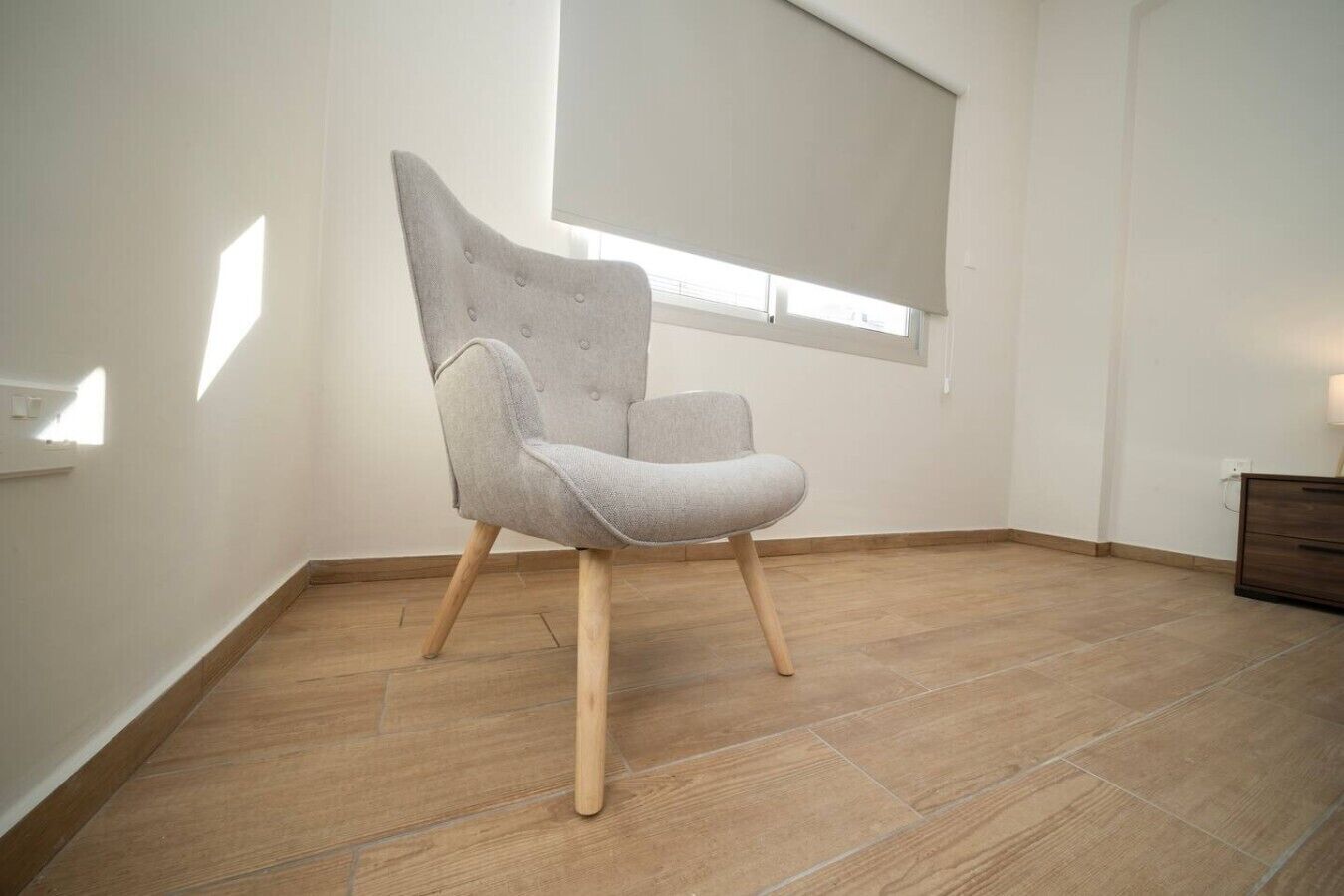 Une pièce avec un fauteuil à revêtement gris, un plancher en bois, une fenêtre avec des stores et une commode en bois.