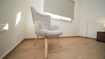 Une pièce avec un fauteuil à revêtement gris, un plancher en bois, une fenêtre avec des stores et une commode en bois.