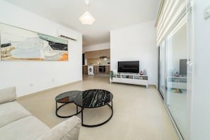 A modern living room with a sofa, two black marble coffee tables, a flat-screen TV on a white entertainment unit, and a kitchen in the background.