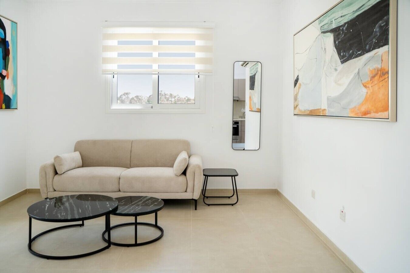 Un salon comprenant un canapé, deux tables rondes, une fenêtre avec des stores, un miroir et un tableau accroché au mur.
