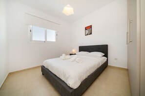 A bedroom with a bed, a nightstand, a wall-mounted light, a window, and a door.