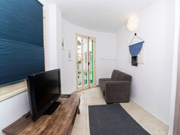 Ein Wohnzimmer mit Fernseher auf einem Holztisch, einem grauen Sofa, einem hängenden Makramee-Sessel und einer Tür, die nach draußen führt.