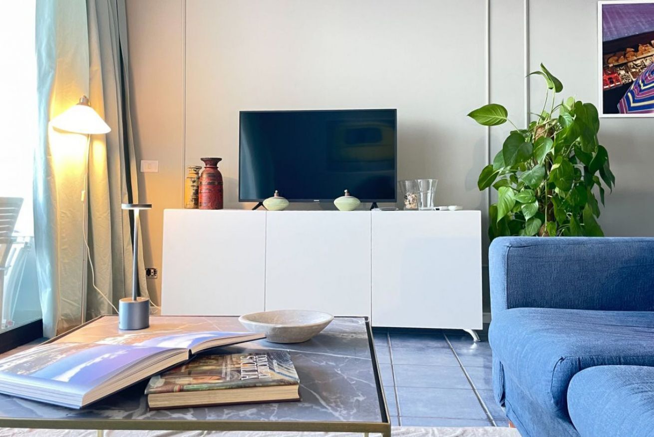 A living room with a flat-screen TV, a sofa, a coffee table with books and a bowl, a lamp, a plant, and a cabinet.