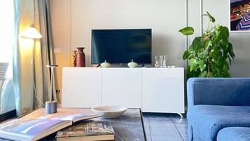 A living room with a flat-screen TV, a sofa, a coffee table with books and a bowl, a lamp, a plant, and a cabinet.