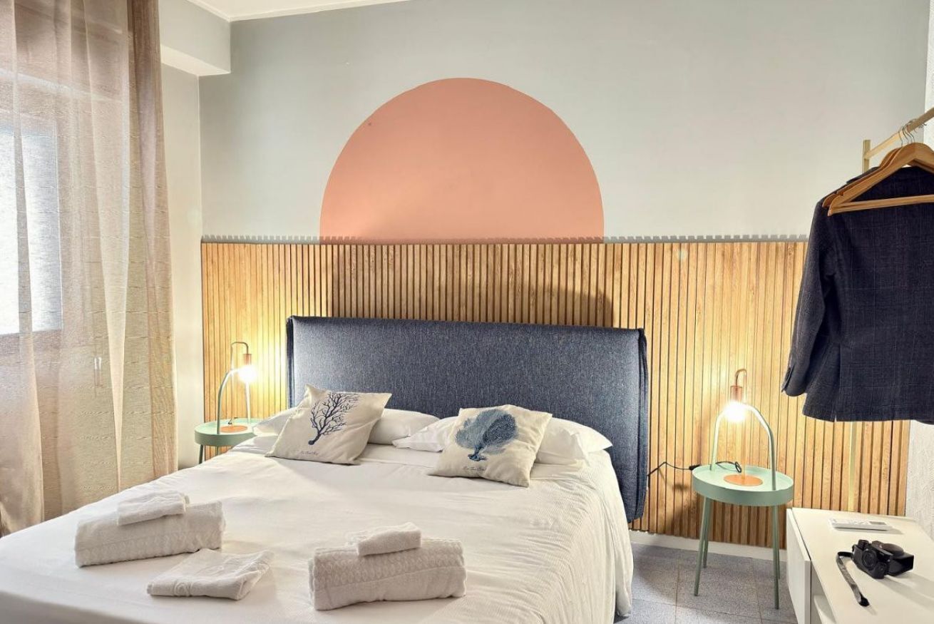 A hotel room with a bed, pillows, a bedside table, a lamp, and a coat rack.