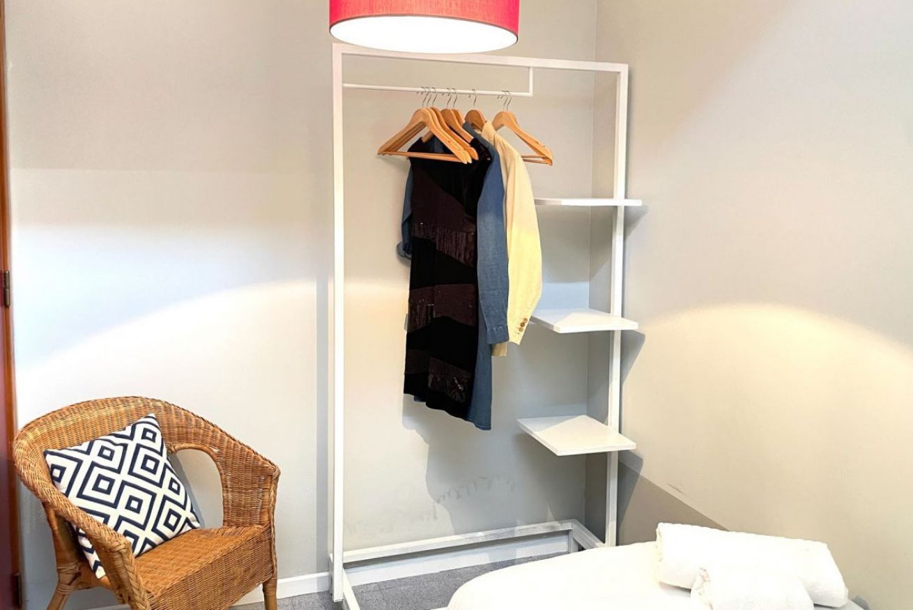 A room with a wicker chair, a hanging coat rack with clothes, and a white shelf unit.
