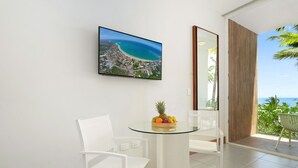 Ein modernes Zimmer mit einem Glastisch, weißen Stühlen, einer Obstschale und einem großen Fenster mit Blick auf Strand und Meer.