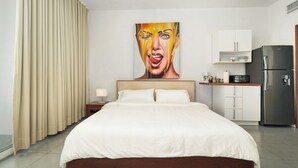 Ein Hotelzimmer mit einem großen Bett, einem Gemälde an der Wand, einer Küchenzeile mit Schränken und einem Kühlschrank.