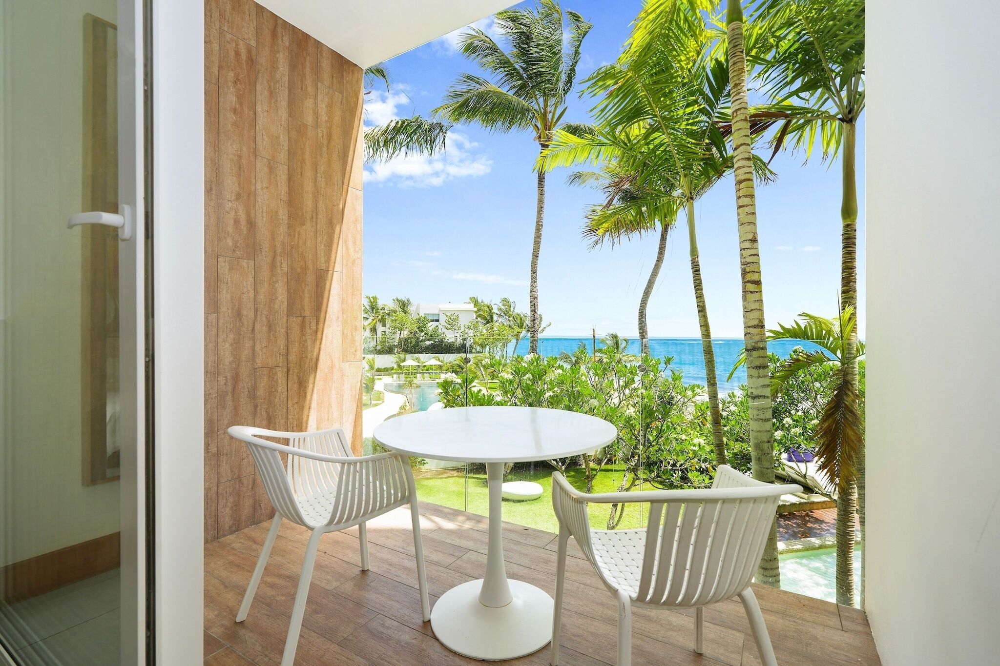 A balcony with a white table and chairs, overlooking a pool and palm trees, with a view of the ocean.