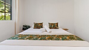 A bedroom with a bed, two patterned pillows, a rolled towel, and a window with a view of greenery. - Fare Manava (Tiahura)