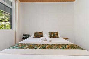 Ein Schlafzimmer mit einem Bett, zwei gemusterten Kissen, einem zusammengerollten Handtuch und einem Fenster mit Blick auf Grünfläche.