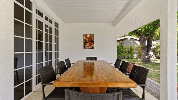 A covered patio with a wooden table and black chairs, large windows, and a potted plant. - Fare Manava (Tiahura)