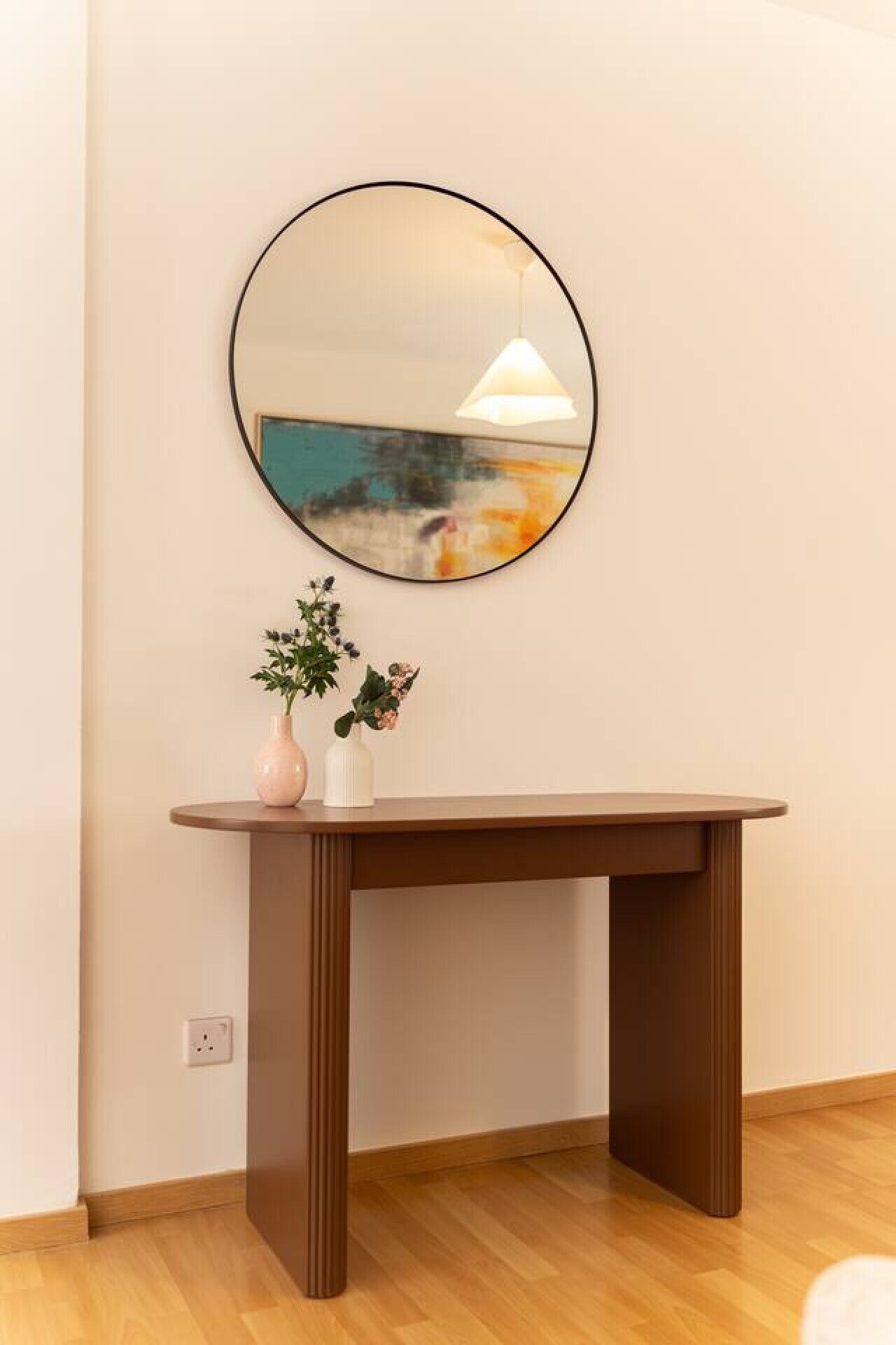 Un miroir rond accroché au mur, une petite table sur laquelle sont posés deux vases, et un luminaire suspendu.