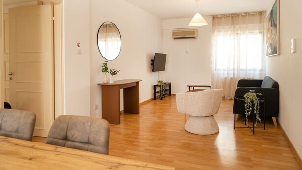 Ein Wohnzimmer mit einer Couch, einem Sessel, einem kleinen Tisch, einem Fernseher und einem Fenster mit Vorhängen.