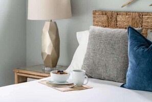 Ein ordentlich gemachtes Bett mit weißer Decke, einem blauen Kissen sowie einer weißen Tasse und einem Untertasse mit einer Schüssel Frühstückscerealien auf einer Unterlage.
