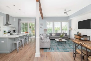 A modern kitchen with bar stools, a dining area with a table and chairs, a sofa, and a TV mounted on the wall. - Walk Everywhere! 12BR Retreat, 3 Pools, Sleeps 24+ (Folly Beach)