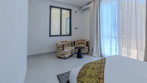 A modern hotel room with a bed, two armchairs, a small table, and a large window with curtains. - 2 or 3 Bedroom - A&E Residence  (Kwale County)