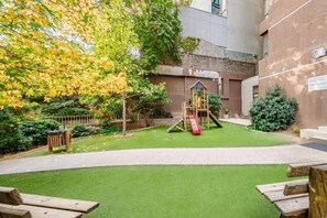Ein Spielplatz mit Rutsche und Klettergerüst, umgeben von einem gepflegten Rasen und einem Gebäude im Hintergrund.