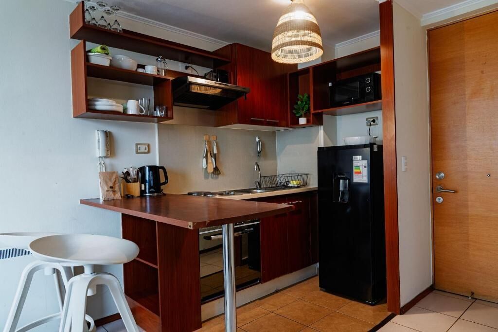 Eine kompakte Küche mit Holzschränken, einem schwarzen Kühlschrank, einer Mikrowelle und einer Pendelleuchte.